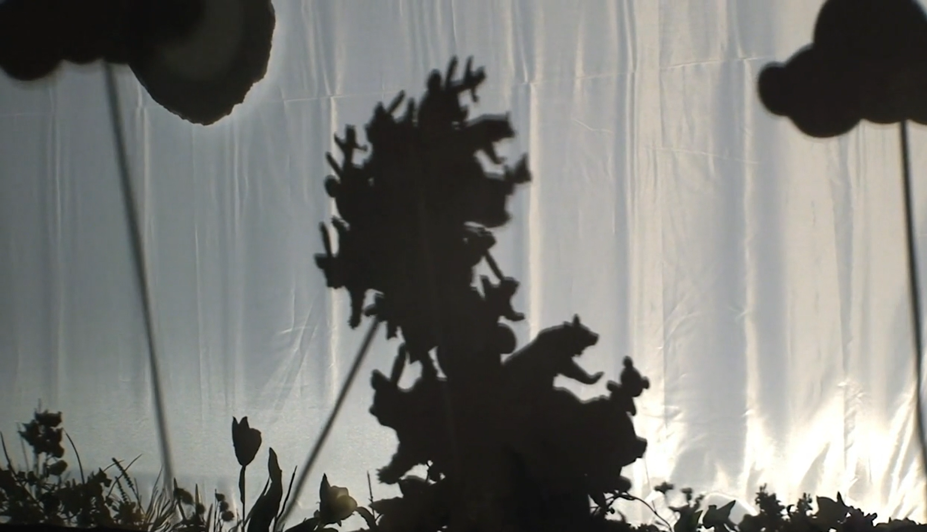 Shadow puppet show with a large tree-like figure, bears, and other animals silhouetted against a bright white screen.